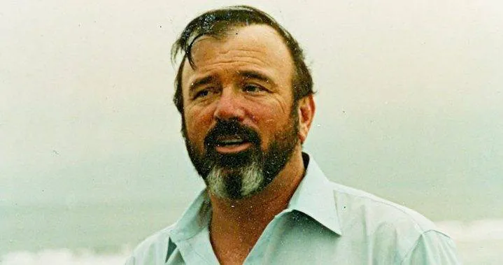 Portrait de Gary Halbert, barbe grisonnante et chemise claire, photographié sur une plage