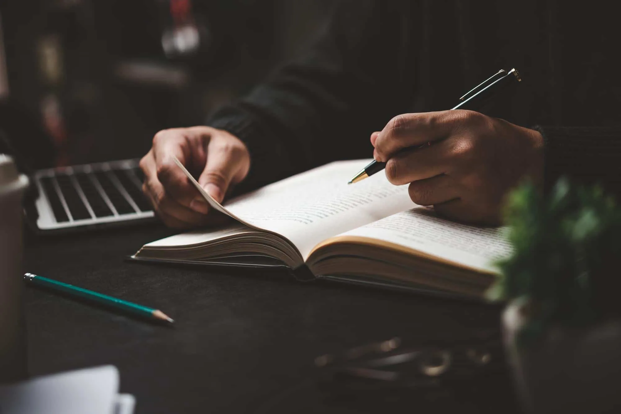 Deux mains tournent la page d'un livre ouvert et l'annotent au stylo, lumière basse